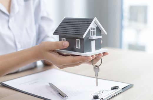 A person holding a miniature home and key on hands. There is also a home loan application and pen on the table.